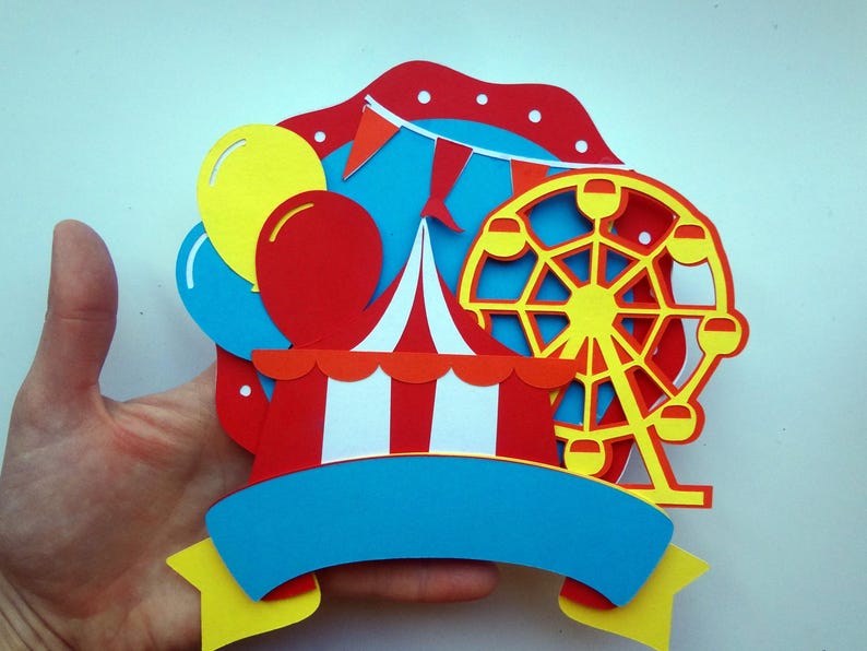 May include: A colorful paper craft featuring a red and white striped circus tent, a yellow Ferris wheel, and blue and yellow balloons. The craft has a blue banner with a yellow border.