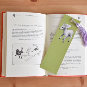 May include: A green bookmark with a black and white illustration of a lamb with two heads. The bookmark has a purple tassel attached to it. The bookmark is open to a page in a book titled "Aesop's Fables" with the story "The Hound and the Hare" on the left page and "The Master Taken Prisoner" on the right page.