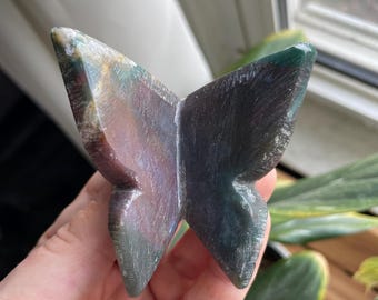 May include: A carved stone butterfly sculpture with a mix of green, purple, and brown hues. The butterfly is held in a hand, showcasing its detailed wing structure and polished finish. The sculpture is a decorative item.