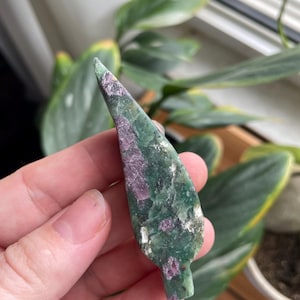 May include: A hand holds a polished, leaf-shaped gemstone. The stone features a mix of green and purple hues, with a textured surface. The gemstone is approximately 3 inches long, and is set against a blurred background of green foliage.