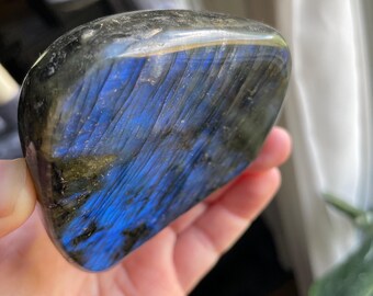 May include: A polished labradorite stone with a mix of dark gray, black, and blue hues. The stone has a smooth, rounded shape and displays iridescent flashes of blue light. This natural gemstone is held in a hand.