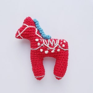 May include: A handcrafted red crochet horse with white and blue detailing. The horse has a blue mane, white decorative stitching, and white dot patterns. This decorative item is a charming addition to any collection.