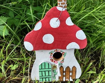 May include: A whimsical, hand-painted birdhouse shaped like a mushroom. The red roof features white polka dots, and the house has a white base with a green door, a brown fence, and a floral wreath around the entry hole. A wire is attached for hanging.