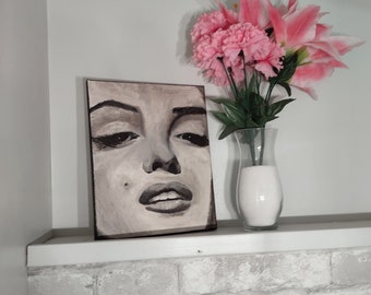 May include: A black and white painting of a woman's face on a canvas, displayed on a white shelf with a vase of pink flowers.