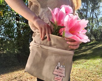 May include: A beige canvas tote bag with a zippered top and a Snoopy doghouse graphic. The bag is filled with large pink flowers. The person is wearing a beige shirt and black leggings.