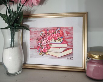 May include: A framed watercolor painting of a stack of books topped with a bouquet of pink roses and other flowers. The painting is in a gold frame. A clear glass vase with white sand and pink flowers is to the left.
