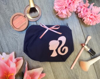 May include: Navy blue cosmetic bag with a pink silhouette of a woman's profile. The bag is decorated with a pink ribbon and is surrounded by makeup items, including blush, brushes, and lipstick. Pink flowers are also present.