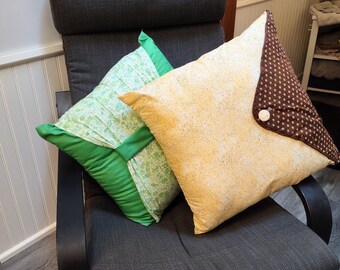 May include: Two decorative throw pillows on a gray chair. One pillow is light green with a floral pattern and green trim. The other is yellow with a speckled design and a brown star-patterned flap. A white button secures the flap.