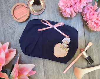 May include: A navy blue cosmetic bag with a pink ribbon bow and a Strawberry Shortcake graphic. Makeup brushes, lipstick, and blush are arranged around the bag, with pink flowers in the background.