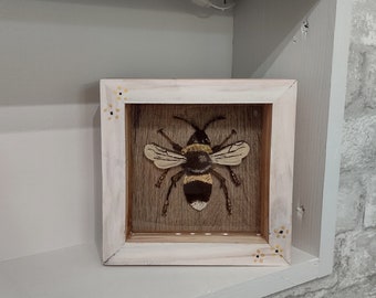 May include: A white wooden frame with a brown background and a painted image of a bee. The bee is black and yellow with white stripes. The frame has a decorative border of yellow dots.