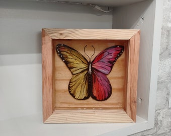 May include: A framed artwork featuring a butterfly with yellow and pink wings. The butterfly is set against a light wood background.
