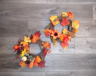 May include: Two autumn wreaths with fall foliage, including orange, red, and yellow leaves, and small white lights. One wreath has a sunflower and berries. The wreaths are made of twigs and are on a wooden surface.