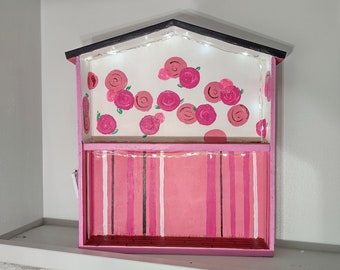 May include: A pink wooden house-shaped shelf with a white background and pink roses painted on it. The shelf has a second level with pink and white stripes. There are white lights strung across the top of the shelf.