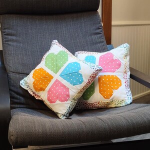 May include: Two square decorative pillows with a patchwork heart design. Each heart is divided into four sections, each a different color: orange, pink, green, and blue, with white polka dots. The pillows are on a gray chair.