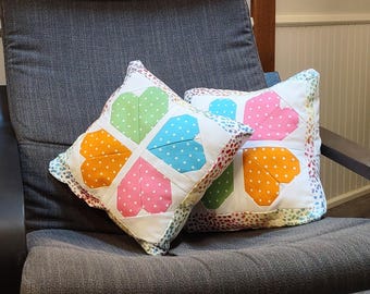 May include: Two square decorative pillows with a patchwork heart design. Each heart is divided into four sections, each a different color: orange, pink, green, and blue, with white polka dots. The pillows are on a gray chair.