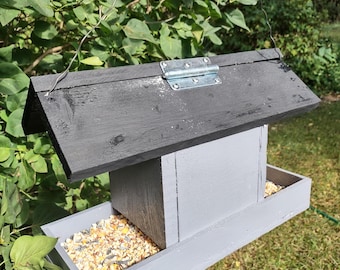 May include: A gray wooden bird feeder with a black roof and a tray filled with birdseed. The feeder is suspended by a wire and is surrounded by green foliage and grass.