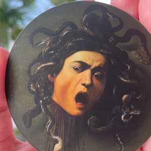 Puede incluir: Una pegatina redonda que presenta un primer plano de la cabeza de Medusa con serpientes como cabello. La imagen es una reproducci&oacute;n de una pintura famosa. El fondo es de color verde oscuro.