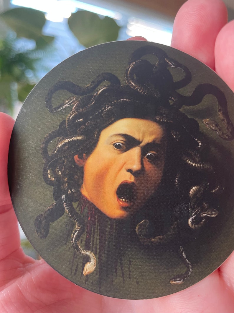 Puede incluir: Una pegatina redonda que presenta un primer plano de la cabeza de Medusa con serpientes como cabello. La imagen es una reproducci&oacute;n de una pintura famosa. El fondo es de color verde oscuro.