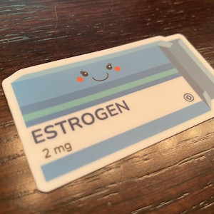 May include: A white sticker with a blue and green pill shape. The pill has a smiling face with eyelashes and red cheeks. The text "ESTROGEN 2 mg" is printed on the pill.