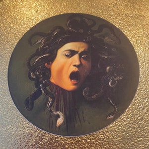 Puede incluir: Una pegatina circular con una pintura de Medusa, una figura mitol&oacute;gica griega con serpientes por cabello, representada con una expresi&oacute;n de horror. El fondo es de color verde oscuro y las serpientes son de color marr&oacute;n oscuro.