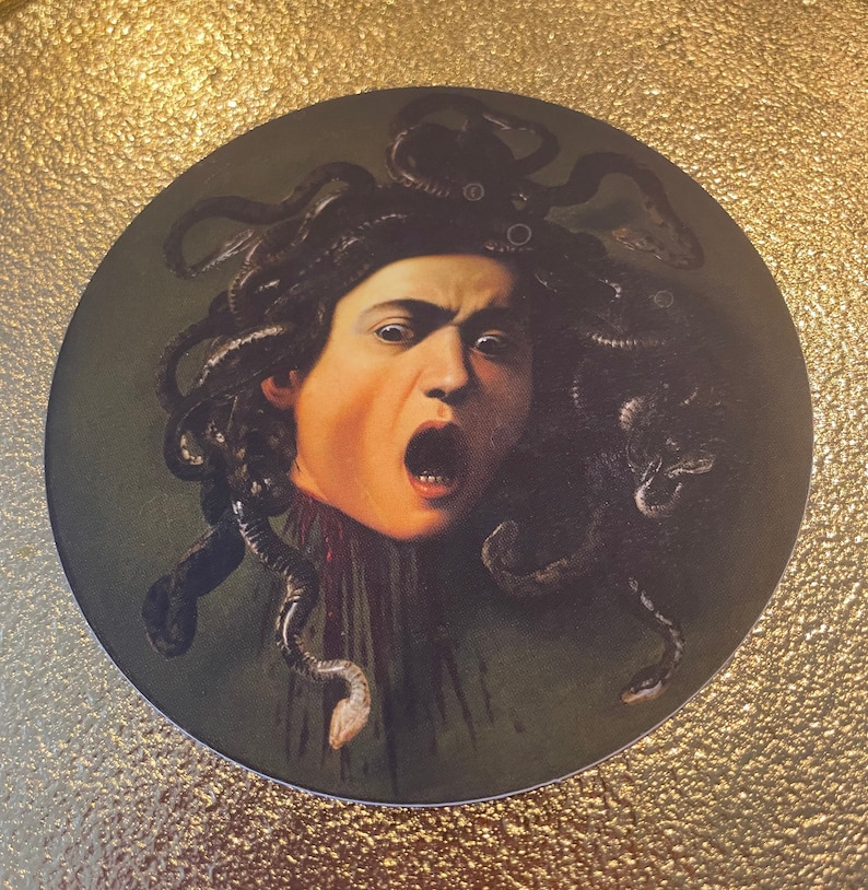 Puede incluir: Una pegatina circular con una pintura de Medusa, una figura mitol&oacute;gica griega con serpientes por cabello, representada con una expresi&oacute;n de horror. El fondo es de color verde oscuro y las serpientes son de color marr&oacute;n oscuro.