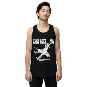 Op de afbeelding: Zwart mouwloos shirt met een witte afbeelding van een cowgirl met een spuit en de tekst "FUCK SAFE SHOOT CLEAN" en "AUSTIN HARM REDUCTION COALITION". Het shirt is mouwloos en gemaakt van zacht materiaal.