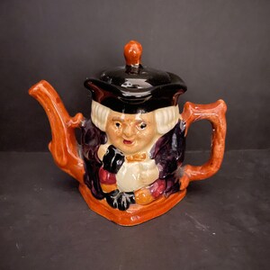 May include: A small ceramic teapot shaped like a man wearing a black hat and a purple robe. The teapot has a brown handle and spout.