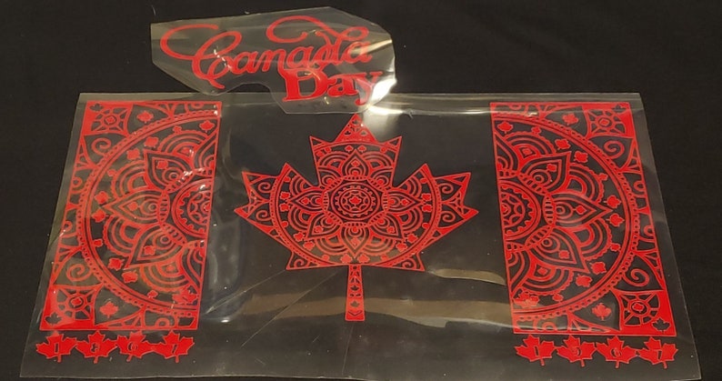 Puede incluir: Calcoman&iacute;a de vinilo rojo de una bandera canadiense con un dise&ntilde;o de mandala. La bandera presenta una gran hoja de arce en el centro y dos dise&ntilde;os de mandala m&aacute;s peque&ntilde;os a cada lado. Las palabras "Canada Day" tambi&eacute;n est&aacute;n incluidas en la imagen.