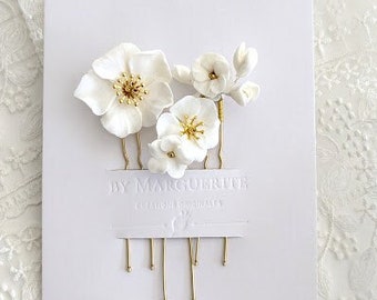 May include: Set of four white porcelain flower hair combs with gold accents. The combs are arranged on a white card with the text "By Marguerite" printed on it.