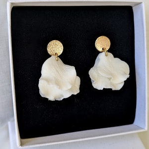 以下が含まれることがあります： 金色のアクセントが付いた白い花の形をしたイヤリングのペア。イヤリングは、「By Marguerite Creations Originales」というテキストが書かれた白い箱に入っています。