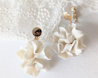 以下が含まれることがあります： オフホワイトの花の形をしたクリップオンイヤリングのペア。各イヤリングは、複数のフリルのある花びらと金色のクリップが特徴です。イヤリングは、白くテクスチャ加工された表面に表示されています。