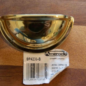 May include: A polished brass semi-circular cup drawer pull with a reflective finish. Attached is a white paper label featuring the Amerock logo, the model number BP4235-B, and a barcode with the text Amerock Corporation, Rockford, IL.