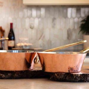 May include: Two copper saucepans with brass handles, sitting on a wooden surface. The saucepans are nested together, with the smaller saucepan inside the larger one.