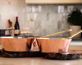 May include: Two copper saucepans with brass handles, sitting on a wooden surface. The saucepans are nested together, with the smaller saucepan inside the larger one.