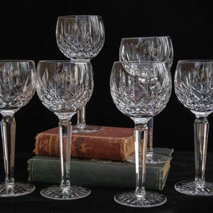 May include: Six clear crystal wine glasses with intricate cut glass patterns. The glasses are arranged on a dark surface, with two antique books in the background. The stemware has a classic, elegant design.