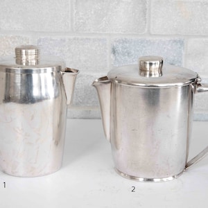 May include: Two silver pitchers with lids and handles. The pitcher on the left is slightly taller and narrower than the pitcher on the right.