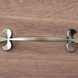 May include: A horizontal metal drawer pull with a brushed brass finish. The handle features a central curved bar flanked by two symmetrical fleur-de-lis motifs at each end, creating a decorative hardware piece for cabinetry or furniture.