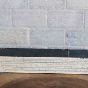May include: A vintage slide rule with a black case and a white face. The slide rule has a silver metal frame and a clear plastic window.