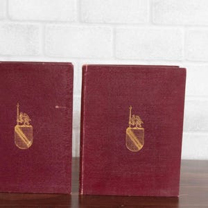 May include: Two burgundy-colored books with a gold emblem on the front cover. The emblem features a shield with a crest and a vertical element. The books are standing upright on a wooden surface.