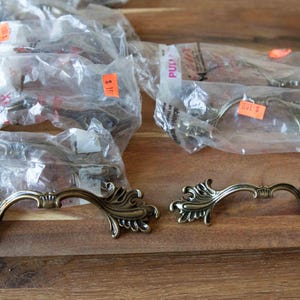 May include: A collection of antique brass-toned metal drawer pulls featuring ornate, scrolling leaf-like designs on each end. Two pulls are displayed unwrapped in the foreground, while several others remain sealed in clear plastic packaging.