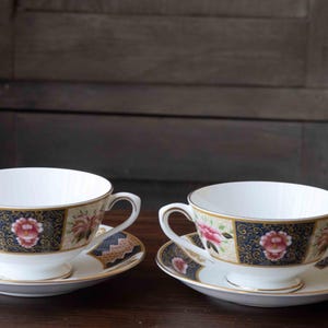 May include: Two white porcelain bouillon cups with two handles and matching saucers. The exterior features alternating panels of dark blue with gold filigree and cream with pink floral patterns, all finished with a thin gold rim on the edges.