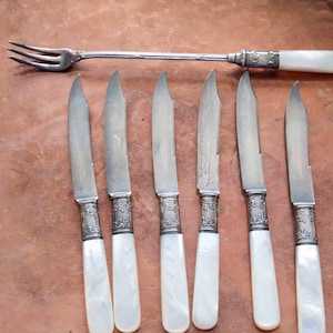 May include: Set of seven silver knives with mother-of-pearl handles. The knives have ornate designs on the blades and handles.
