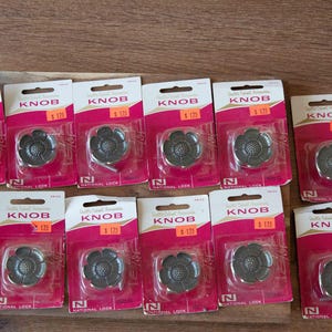 May include: A collection of twelve individual metal cabinet knobs, each shaped like a flower with a textured center. Each knob is sealed in its own white and magenta retail packaging labeled with the word KNOB and the National Lock logo.