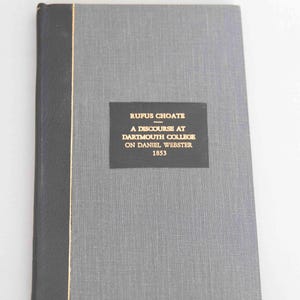 May include: A hardcover book with a grey textured cover and a black spine. A black rectangular label on the front cover features gold text that reads: RUFUS CHOATE - A DISCOURSE AT DARTMOUTH COLLEGE ON DANIEL WEBSTER 1853. The spine is black.
