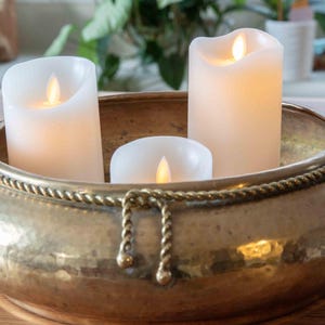 May include: A round, hammered brass bowl featuring a decorative rope-style trim with a hanging knot detail. Inside the bowl are three white, cylindrical LED pillar candles with warm, glowing flames, creating a soft, ambient light effect.