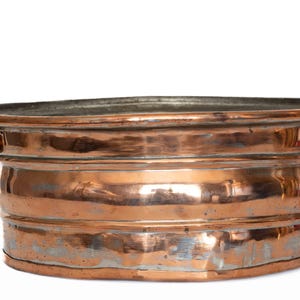 May include: A copper-colored oval tub with brass handles. The tub has a wide rim and a series of horizontal bands around its body. The metal has a reflective surface with some areas of discoloration, suggesting age or use.