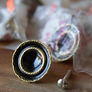 May include: A round cabinet knob featuring a glossy black center, a gold-toned rope-textured inner ring, and a scalloped gold-toned outer edge. The knob includes a silver-toned metal mounting screw positioned next to it on the surface.