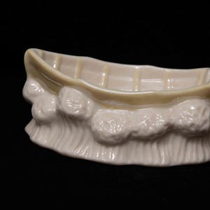 May include: A cream-colored ceramic boat figurine featuring a smooth, open hull design with vertical interior ribs. The exterior of the vessel is decorated with a sculpted, textured wave pattern that encircles the base, creating a relief effect.
