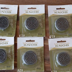 May include: Six individual packages of National Lock cabinet knobs. Each package contains one round, dark metal knob with a central cross design and a rope-like border. The packaging is olive green and white with the text Quality Cabinet Accessories KNOB.