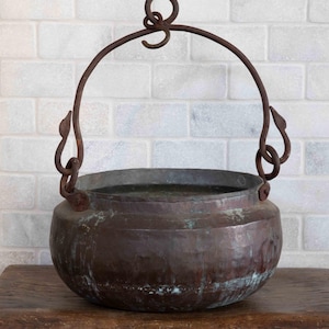 May include: A rustic, antique copper cauldron with a curved handle and decorative side attachments. The pot sits on a wooden surface against a white brick wall. The cauldron's aged patina gives it a vintage aesthetic.